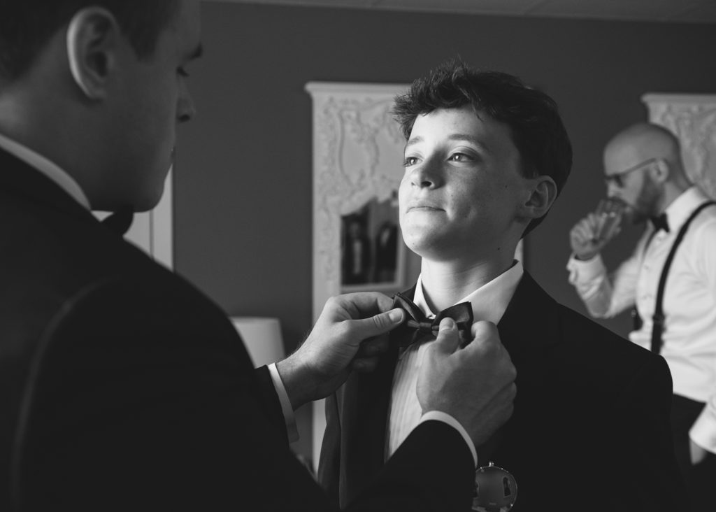 Black and white photograph of older brother helping younger with his tie on morning of the wedding.