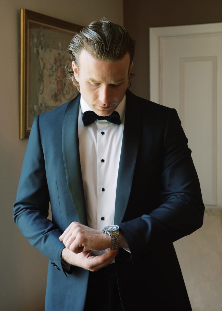 Groom dressed in tuxedo in morning of wedding while he fixes his watch.