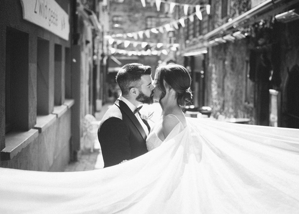Black and white portrait of Galway wedding couple sharing kiss with dress veil cape in foreground.