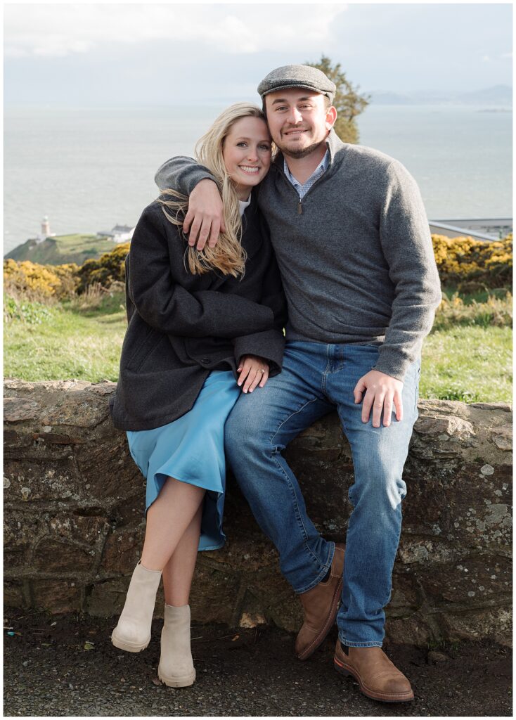 Engaged couple Emily and Jacob hug on the top of Howth Summit, Dublin, with The Baily Lighthouse behind them.