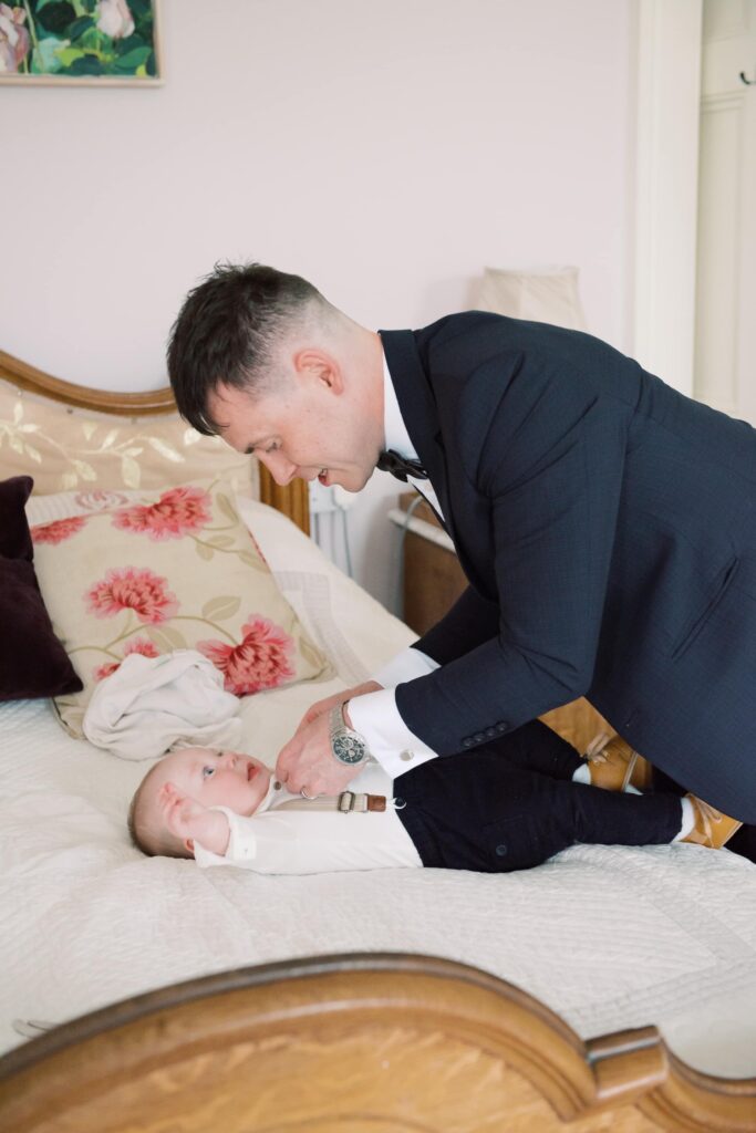 Groom dresses his giggling baby son in his cute wedding day outfit.