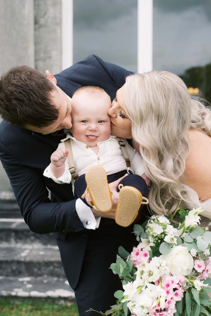 Adorable portrait of the family: bride, groom, and their baby son being kissed on both cheeks!