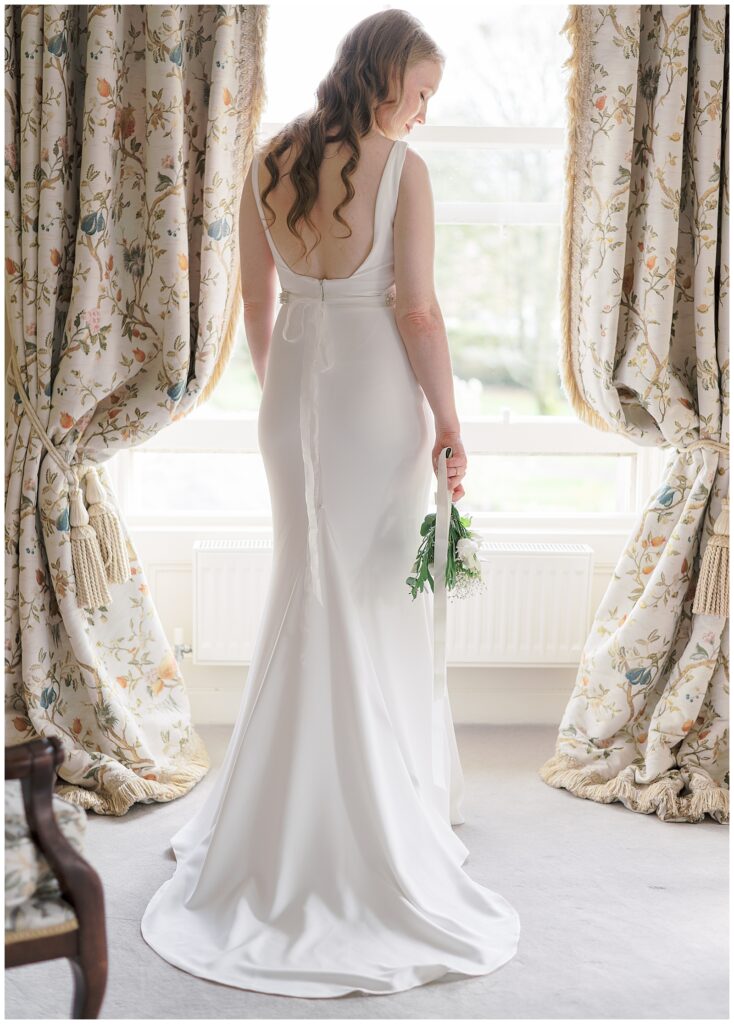 Portrait of the bride's back as she stands in front of a big bright window with her bouquet held down by her side.