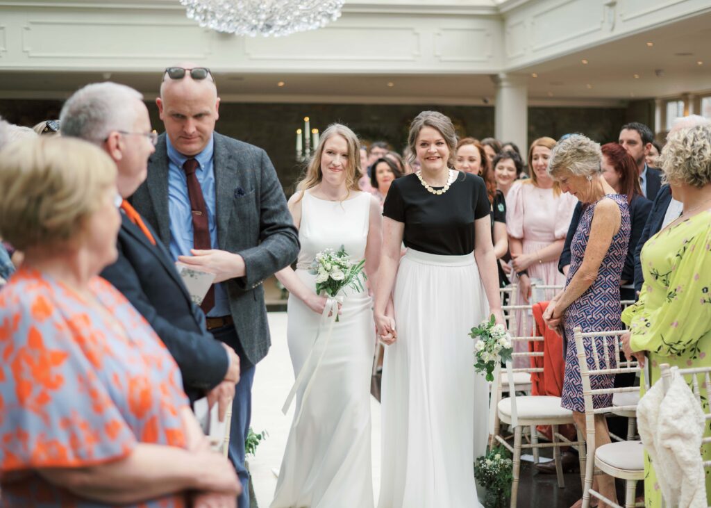 Brides walk down the aisle together for their same-sex wedding ceremony in the Orangery.