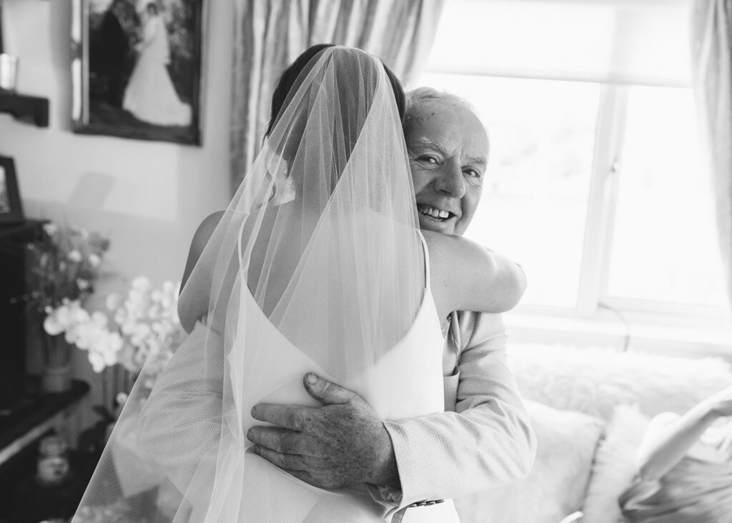 Black and white portrait of the bride and her father sharing a hug in the family home.