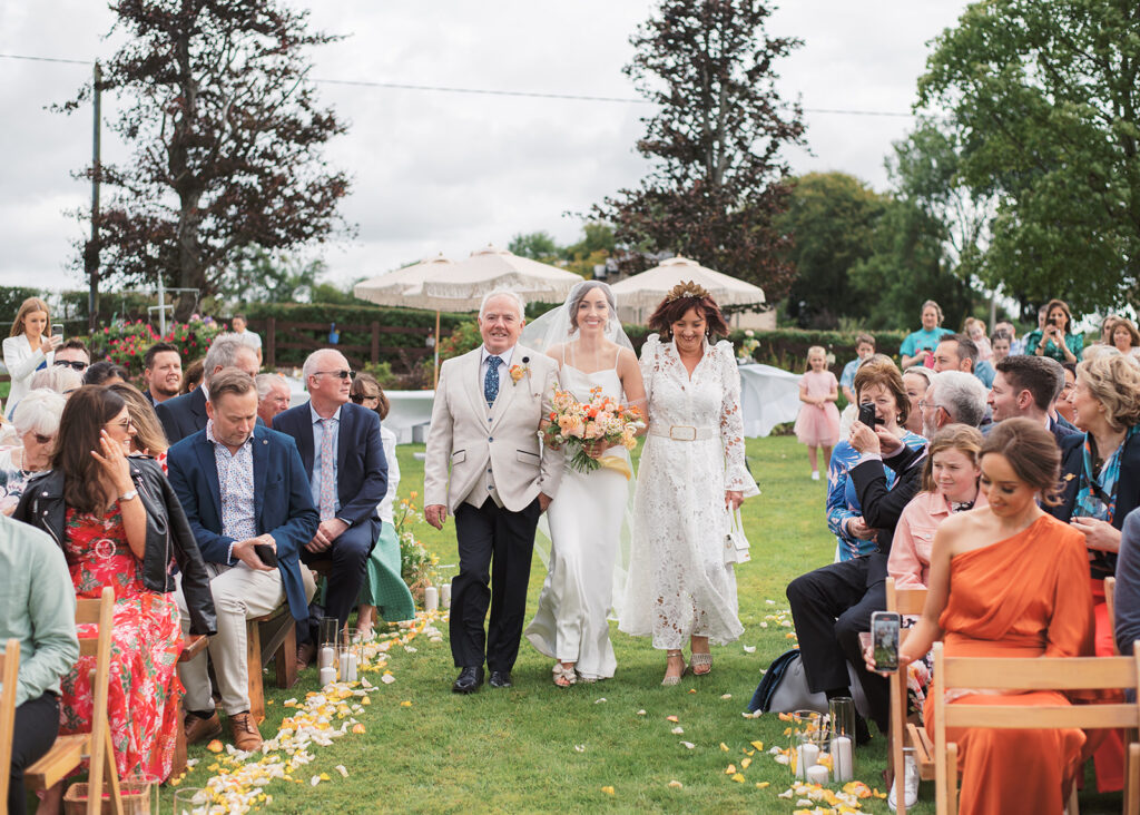 Bride and her parents walk together down the aisle of their garden wedding ceremony.