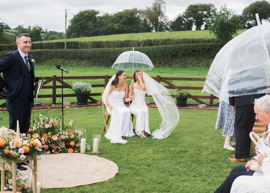 The brides hide under an umbrella together during their outdoor wedding ceremony.