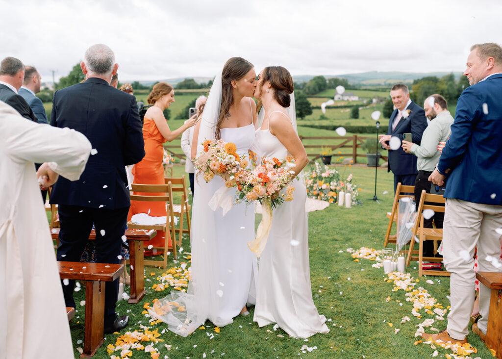 Two brides share a kiss in the aisle after their outdoor wedding ceremony and are showered with confetti and flower petals.